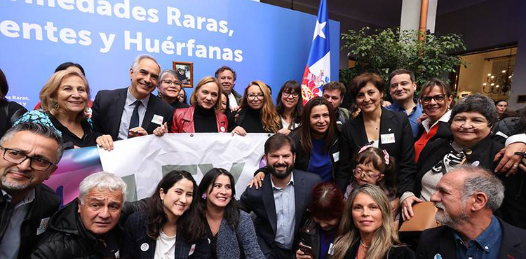 Grupo de autoridades, familias y organizaciones reunidas en la ceremonia de promulgación de la Ley de Enfermedades Poco Frecuentes en Chile, junto a la bandera nacional